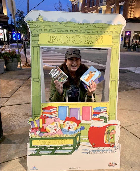 A woman in a hat and coat holding up two books inside a cardboard cutout with pictures of Richard Scarry characters in a sled that says "Shop Local"