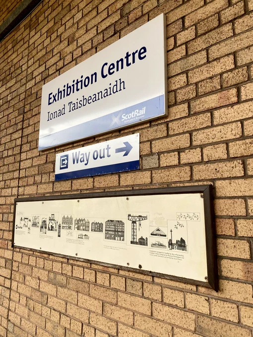 This is the sign for Exhibition Centre Station. Below it is the explanatory plaque for Glasgow artist Willie Rodger’s 1988 mural. Both are on a yellow brick wall.