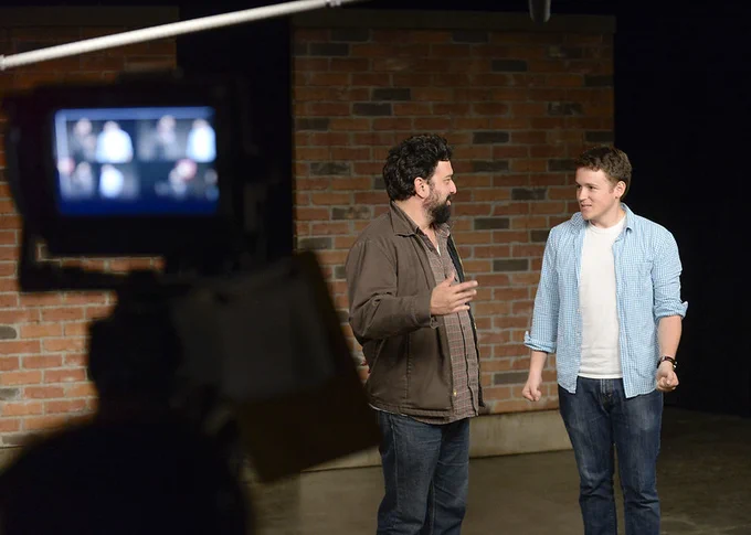 Behind the scenes, a cameraman films two men in casual attire against a brick wall, one in a brown jacket and the other in a blue checkered shirt, seemingly in discussion.