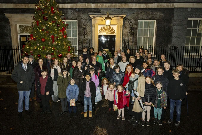  A large group of adults and children stand outside the door of 10 Downing Street with a Christmas tree behind them on the lefthand side.