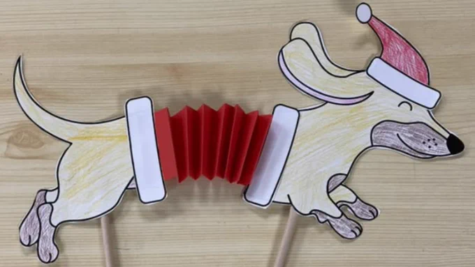Handcrafted paper cut-out of a dachshund wearing a Santa hat, with a red accordion-style body, displayed on a wooden surface.