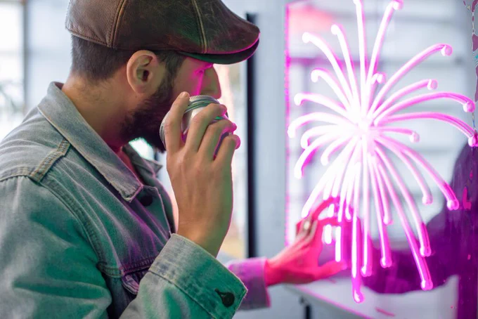 Artist Carmen Papalia holds an audio device to his ear while touching a pink-lit acrylic panel illuminated with a dahlia firework with arching trails. 