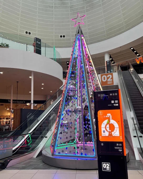 A vibrant Christmas tree made of lights and reflective ornaments displayed in a shopping mall atrium, topped with a pink star.