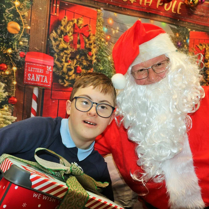 Boy holding a wrapped Christmas gift beside Santa Claus in a festive setting.
