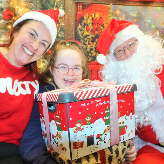 Santa Claus posing with woman and child holding a festive gift box decorated with Christmas characters.