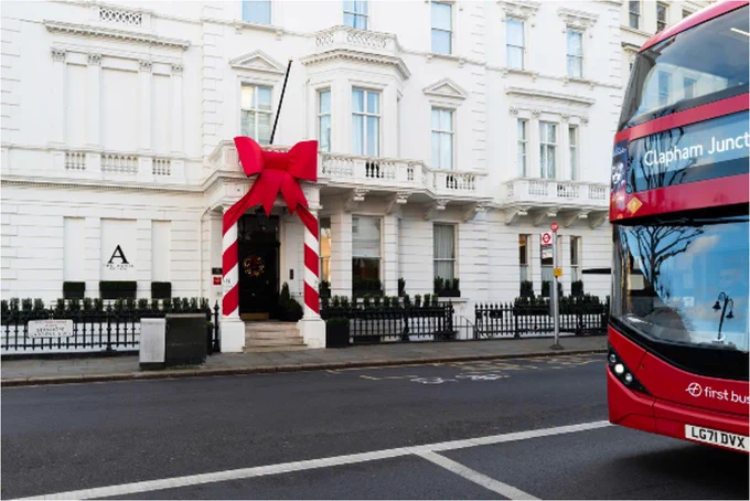 The front of a red London bus, driving along a road with white houses and a front door with a red bow