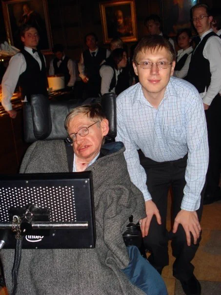 Dr. Mihran Vardanyan with Professor Stephen Hawking at a Cambridge University formal dinner. Hawking is in his wheelchair with his speech synthesizer, and traditional Cambridge oil portraits are visible in the background.