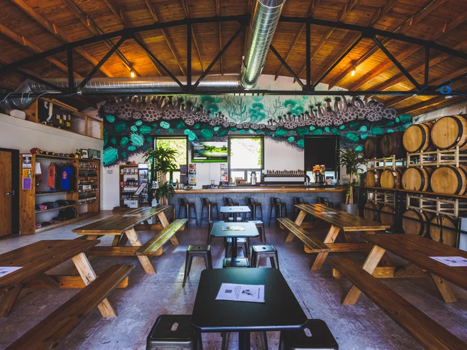 Interior of Speciation Cellars with wooden picnic tables, a bar at the end, and a colorful mural on the wall.