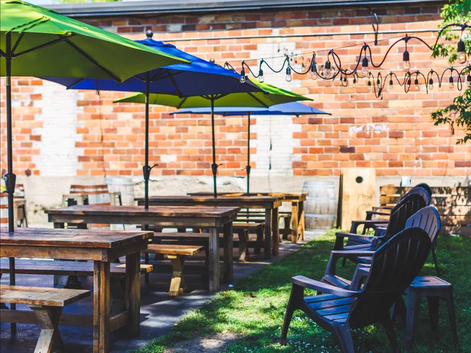 Outdoor cafe patio with colorful umbrellas, wooden picnic tables, and modern chairs, set against a brick wall with decorative lights.