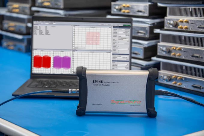 Signal Hound SP145 spectrum analyzer connected by cables on a workbench, with analytical software open on a laptop and multiple server racks in the background.