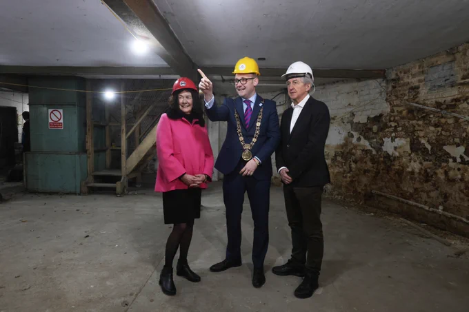 Three people in construction helmets stand in the basement of a tower otherwise known as St. Patrick's Tower. The central figure, wearing a mayoral chain, gestures upwards while the others watch.