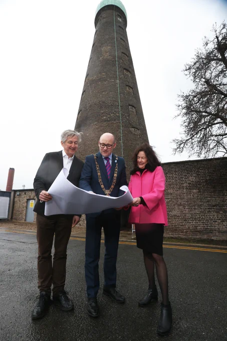 Three people examine blueprints in front of a tall, narrow brick tower otherwise known as St. Patrick's Tower. The central figure wears a ceremonial chain. There is a pear tree peeking out to the right of the image.