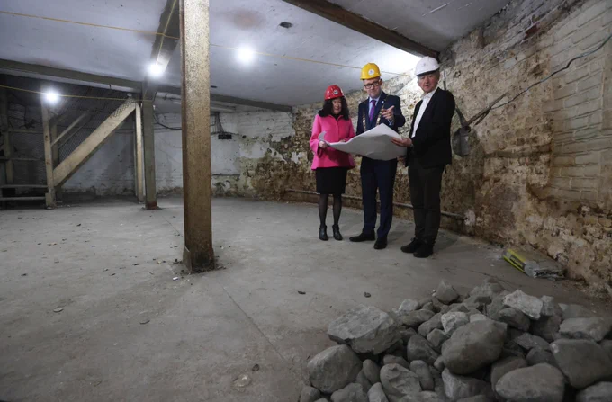 Three people in construction helmets examine blueprints in a dimly lit basement with rough walls and a pile of rocks in the corner.