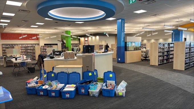 Interior of a public library with bright lighting, featuring rows of bookshelves, a central circulation desk, and several blue bins of books and materials in the foreground.