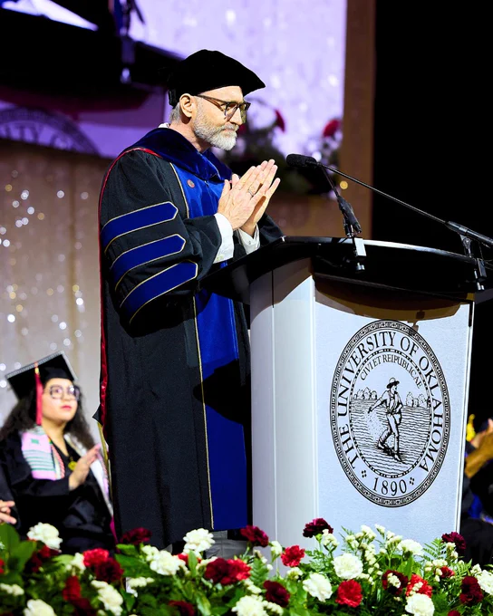A person in academic regalia stands at a podium with the University of Oklahoma seal, applauding at a graduation ceremony, surrounded by floral arrangements.