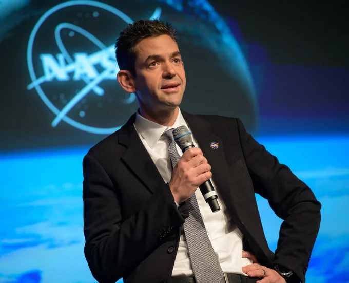 NASA Administrator Jared Isaacman speaking into a microphone at a town hall event. He is wearing a black suit jacket, a white dress shirt, and a textured silver tie, accented with a blue NASA lapel pin and a black wristwatch. The background features a large screen displaying the NASA insignia, illuminated with vibrant blue lighting that contrasts with his formal attire.

Credit: Bill Ingalls