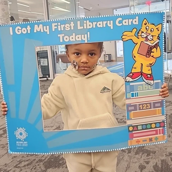 A child wearing a beige outfit holds a large sign that reads "I Got My First Library Card Today!" The sign features a cartoon cat and colorful books. The setting appears to be inside a library, with shelves and bright lighting in the background.