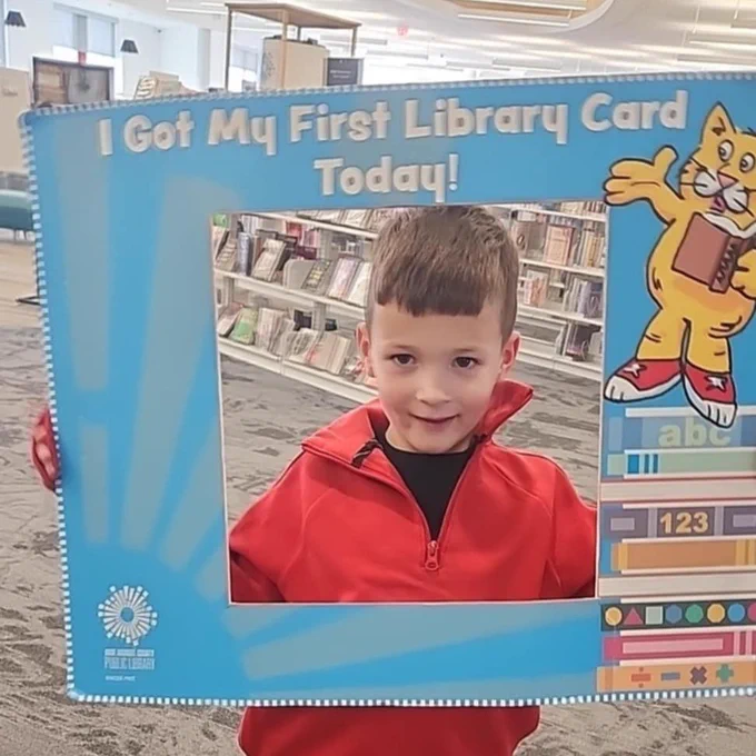 A young boy smiles while holding a colorful frame that reads, "I Got My First Library Card Today!" He is wearing a red jacket and stands in a library setting filled with bookshelves.