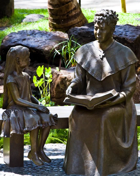 Bronze statue of Bernice Pauahi Bishop reading a book to a young child, situated in a garden with rocks and greenery in the background.