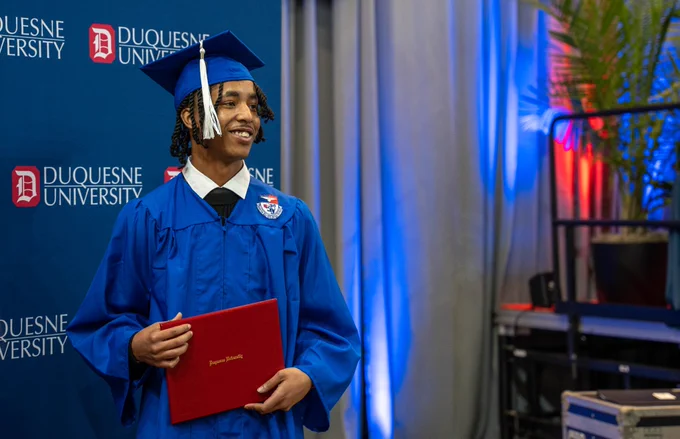 Graduate stands with diploma in front of step and repeat.