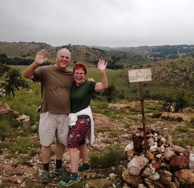 Two parkrunners, with arms around each other's shoulders, waving at the camera next to a 'parkrun' sign in a hilly landscape.