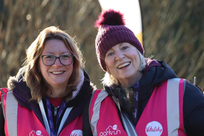 Two parkrun volunteers, one wearing a red beanie, smiling joyfully on a sunny day.