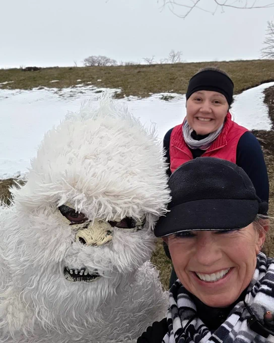 Two parkrunners, one in a pink vest and the other wearing a black cap, smiling and taking a selfie with another parkrunner dressing as a yeti with snow in the background. 