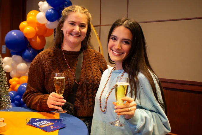 Two individuals smiling and holding glasses, standing at a celebration event with balloons in the background.