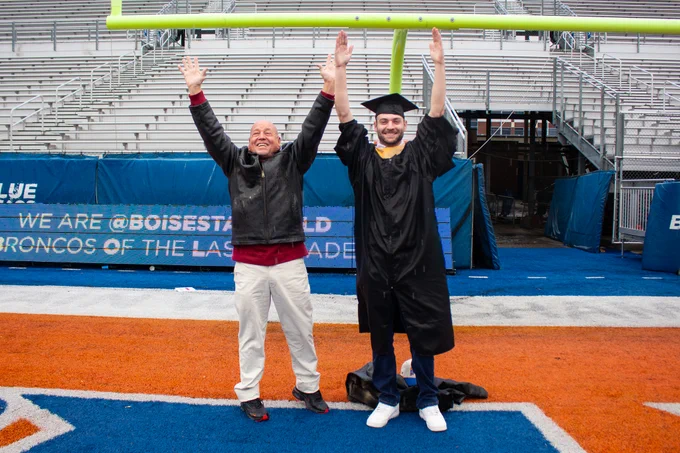 Two individuals celebrate on Boise State University's blue football field; one is in graduation attire and the other in casual clothing, both raising their hands joyously.