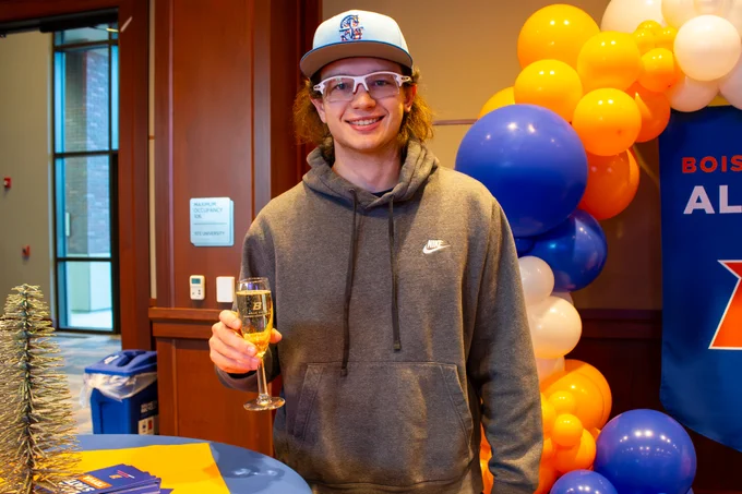 A person in a Boise State University cap smiling and holding a champagne glass, standing in front of a table with blue and orange balloons and decorations.
