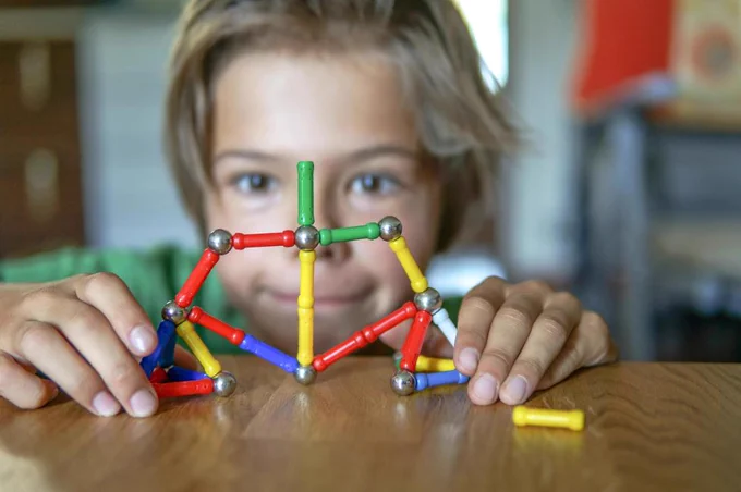 A child peers over a colorful geometric structure made with magnetic building sticks and balls on a wooden table.