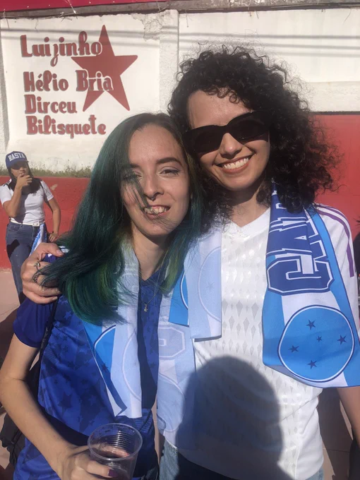 foto nossa tirada pelo thiago, ela me abraça fortemente de lado, estou segurando um copo, estamos sorrindo, as duas vestidas de cruzeiro e com uma faixa das cabulosas no ombro. rapha está de óculos de sol. estamos em frente ao estádio.