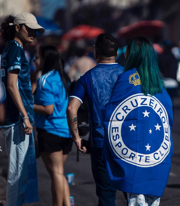 eu e a rapha conversando enquanto caminhamos na rua antes do jogo. ela está na calçada de frente pra mim, com roupa do cruzeiro e de boné. eu estou ao lado dela, andando pra frente, com uma bandeira do cruzeiro e conversando com ela.