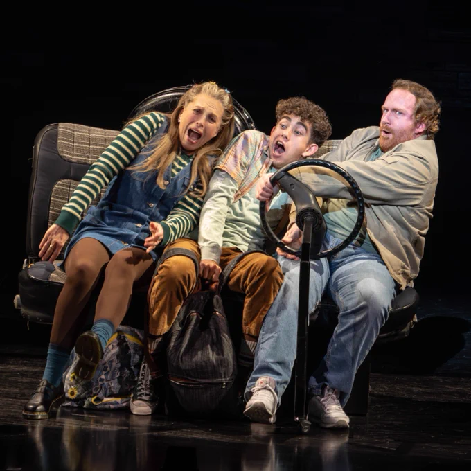 Three actors on stage, one holding a steering wheel, express shock and concern during a scene from a theater production.