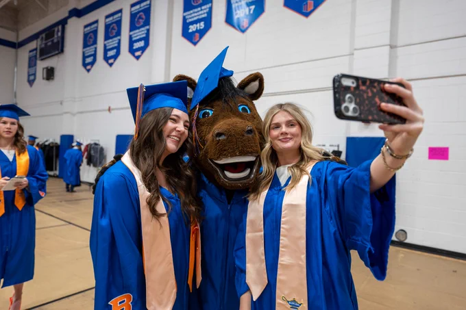 Two Boise State graduates taking a selfie with Buster Bronco wearing graduation regalia. 
