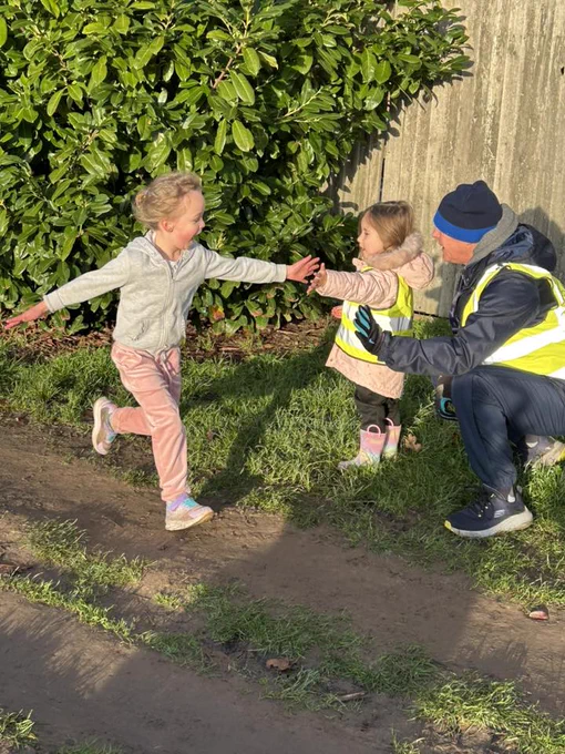 A junior parkrunner and a little volunteer on the left, who is held by an adult kneeling down, making high-five at a parkrun event.