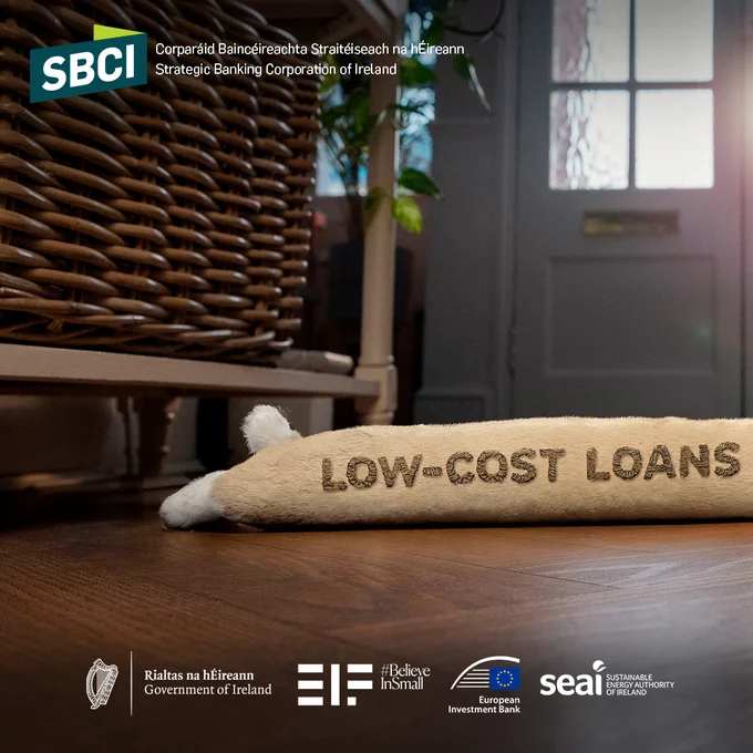 Promotional image for SBCI featuring a plush bolster with 'Low-Cost Loans' written on it, placed on a wooden floor. In the background, there's a wicker chair and elegantly dim lighting. Logos of Rialtas na hÉireann, EIF, European Investment Bank, and SEAI are visible at the bottom.