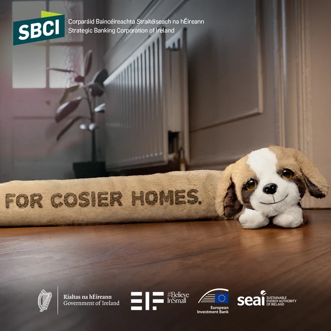 A plush toy dog lies next to a tan door draft stopper with the text "FOR COSIER HOMES" on a wooden floor. The image includes logos for Strategic Banking Corporation of Ireland, Government of Ireland, European Investment Bank, and SEAI at the bottom.