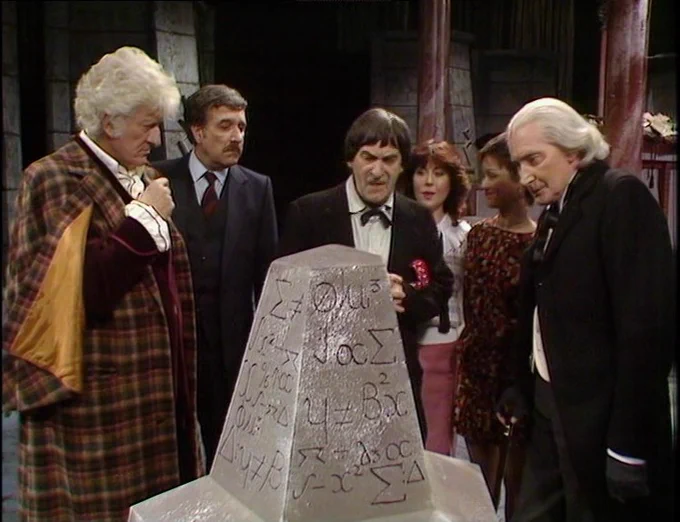 The first three Doctors are gathered around a polystyrene plinth covered with haphazardly deployed mathematical symbols. They are deep in thought. Behind them we can see the Brigadier, Sarah and Tegan.
