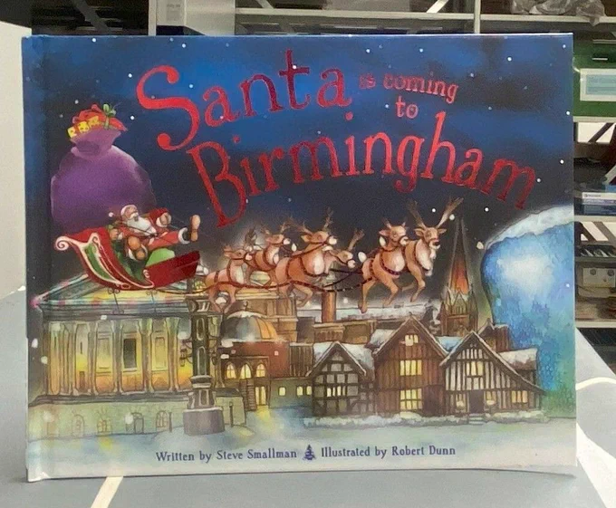 A book on a flat surface. Front cover of the book is facing on to the camera. Title of the book is Santa is Coming to Birmingham. Colourful illustration of Santa on a sleigh above iconic buildings of the city including the Town Hall on the left and Selfridges on the right. Behind the book is shelving.