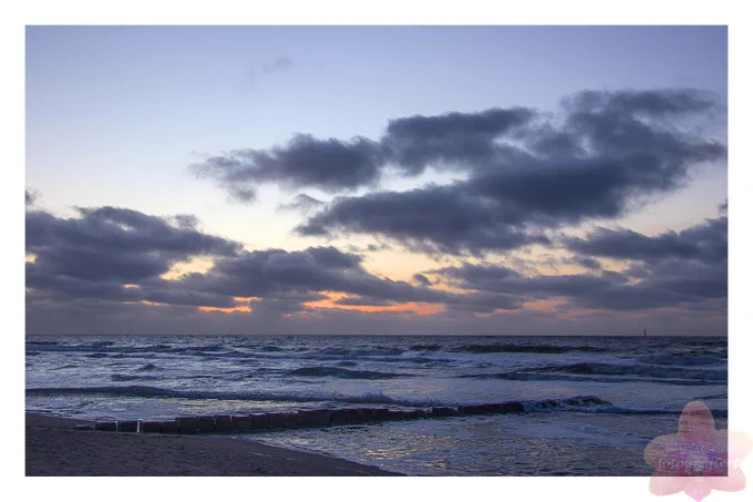 Wolken vor orange Sonnenuntergang und ein Stückchen Strand und schäumende Wellen bis zum Horizont