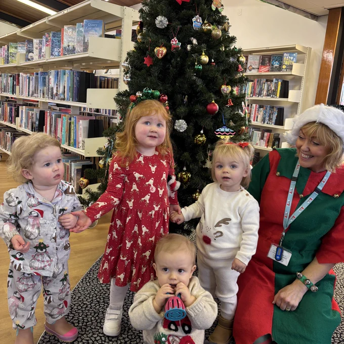 Young children and librarian Cathy, dressed as an elf, stand in front of a Christmas tree. In the background are bookshelves.