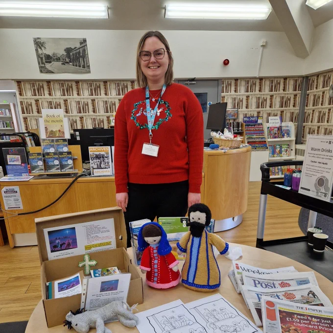 Knitted representations of Mary and Joseph are placed on a table in Freckleton Library. Library assistant stood behind the table smiling. 