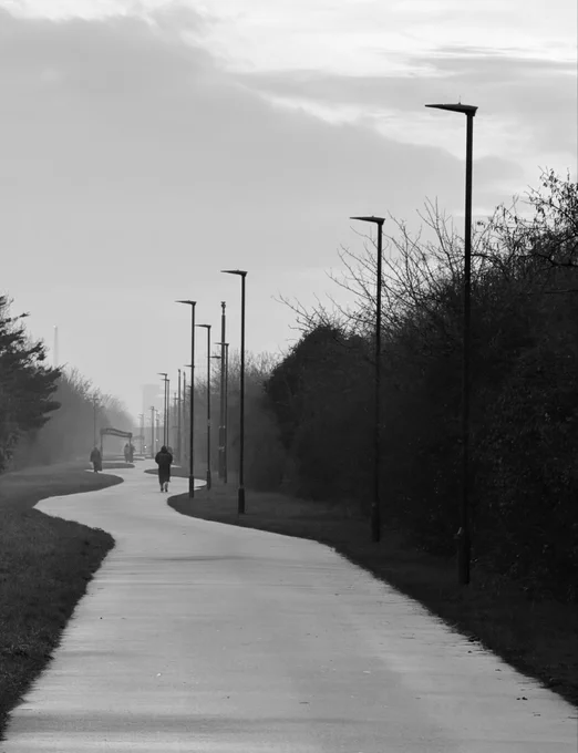 Black and white scene of a path in the mist. Silhouettes of people walking and street lights are visible.