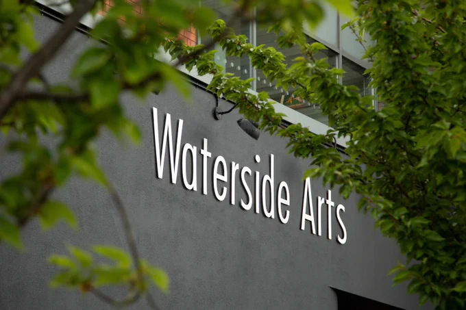 White Waterside Arts sign mounted on a gray building wall partially obscured by green tree branches.