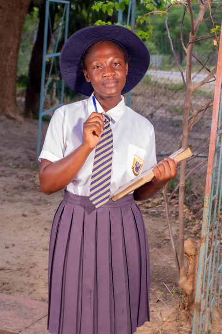 Student in school uniform holding a pen and work book, looking to camera kindly. 