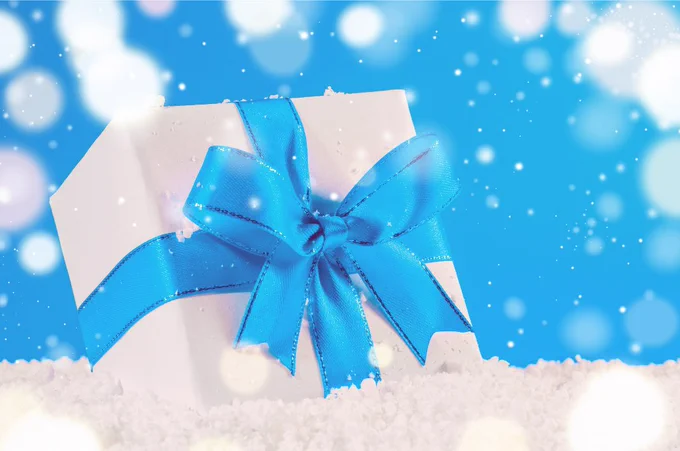 White gift box wrapped with a shiny blue ribbon bow resting on artificial snow against a bright blue background.