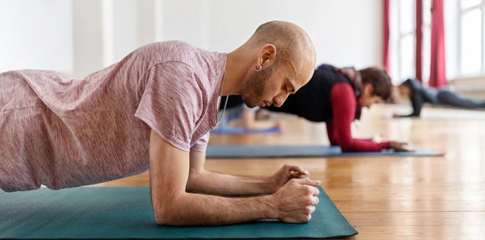Person practicing a plank position on a yoga mat in a bright studio with others in the background