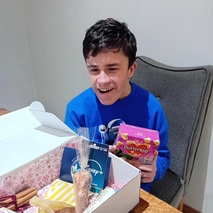 Boy in blue sweater holding a pink Butterkist popcorn box while sitting at a table with an open gift box containing snacks and a Disney  card.