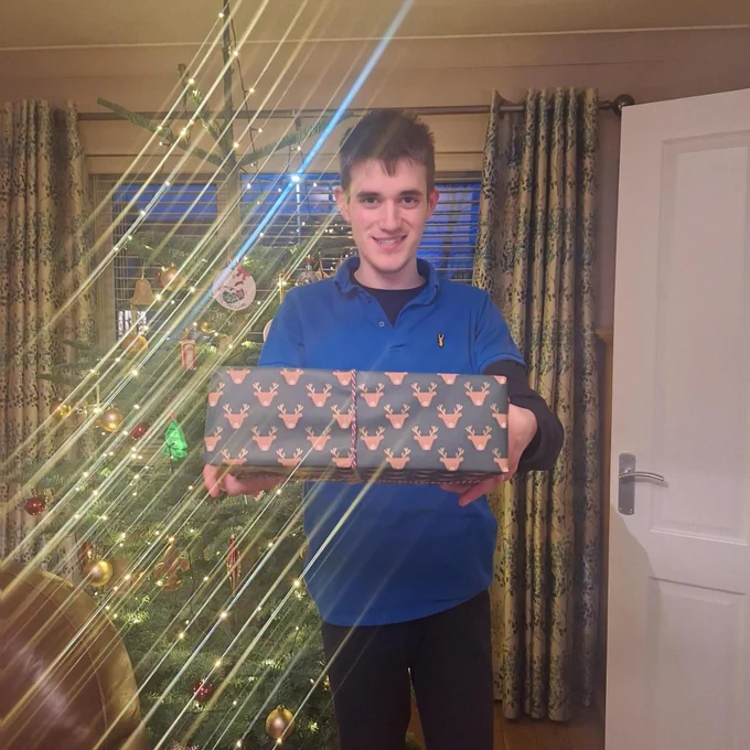 Person in blue shirt holding two reindeer-patterned Christmas presents in front of a decorated tree with lights.
